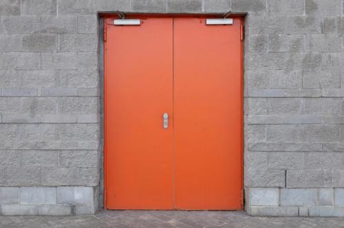 Gray brick wall with wide bright orange door in the centre of the photo. Entry into building